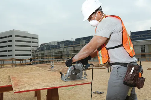 Person wearing safety gear, using a circular saw on a construction site.