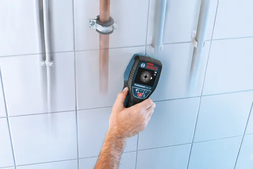 Bosch Professional D-tect 120, a hand-held wall scanner, held against white tiled wall, next to copper and white pipes.