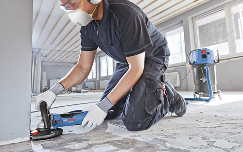 A person wearing safety gear kneels on a floor, operating a Bosch CSG 15 grinder; a blue and black vacuum cleaner is to the right.