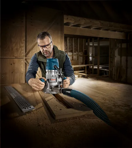 A person wearing safety glasses works with a Bosch router on a wooden surface, connected to a hose.