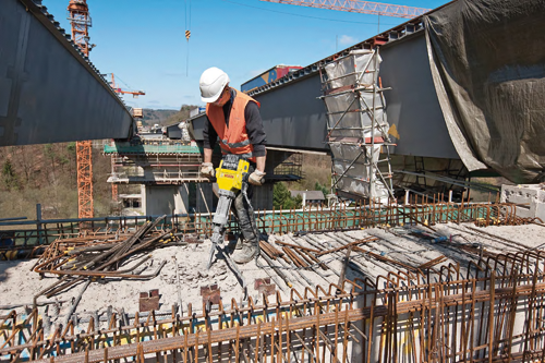 Person wearing a white hard hat and orange vest, operating a yellow and black "BRUTE" Bosch jackhammer on a construction site.