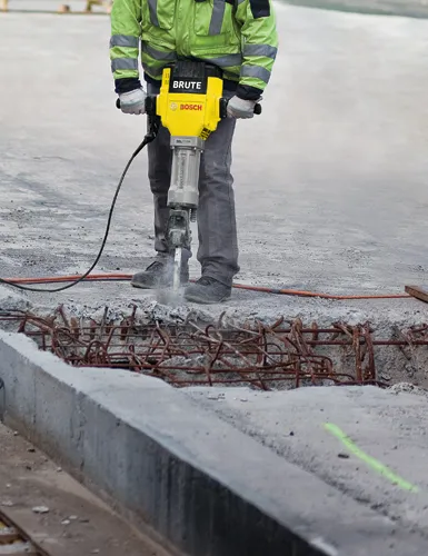 Person using a yellow and black Bosch Brute hammer drill to break up concrete. The concrete has exposed rebar.