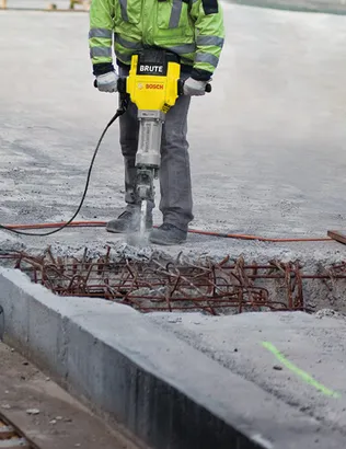 Person using a yellow and black Bosch Brute hammer drill to break up concrete. The concrete has exposed rebar.