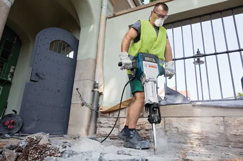 A person wearing protective gear operates a "Brute Turbo" Bosch Boschhammer tool, breaking concrete. The person wears a yellow vest, gloves, and a face mask.