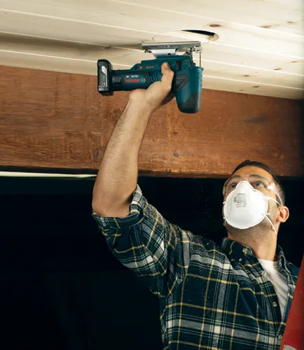 Bosch JS120 HD jigsaw cutting a hole in a wooden ceiling, worker wearing safety glasses and a mask.
