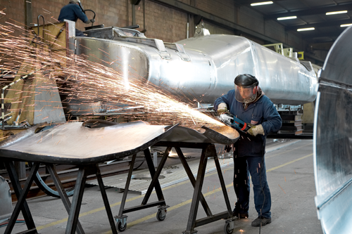 Person using an electric grinder on a metal sheet, sparks flying.  Another person is working in the background.