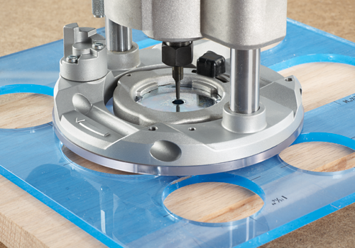 A woodworking router in use with a blue transparent guide plate, positioned on top of a wooden board. The router has a silver finish, and a bit extends downward through a hole in the guide plate and into the wood.