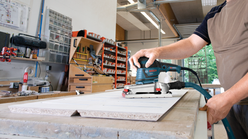 Man uses Bosch OS50VC orbital sander on white surface in a workshop.