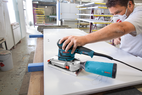 Person wearing a mask uses a Bosch OS50VC sander to sand a white surface.