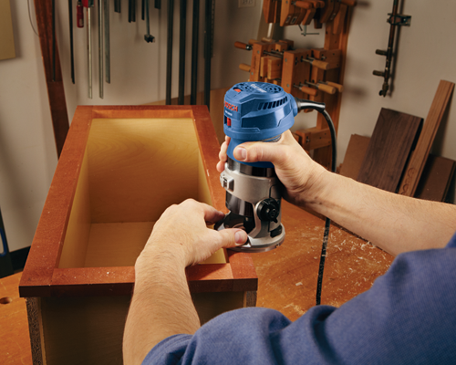 Bosch GKF125CE, a blue and silver router, being used on a wooden cabinet by a person.