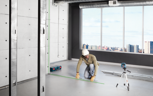 Person wearing hard hat and gloves working with a green laser level and metal studs in a construction setting.