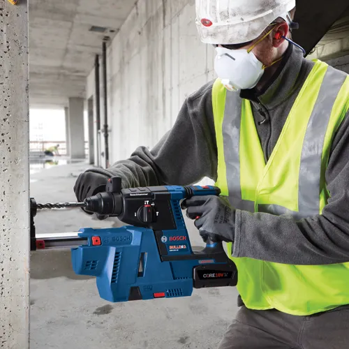 A person wearing a hard hat, safety glasses, mask, earplugs, high-visibility vest, and work gloves drills a hole in a concrete pillar with a blue and black Bosch BULLDOG GBH18V-26 cordless rotary hammer drill; the drill has a CORE 18V battery.