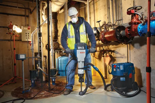 Person wearing a hard hat, safety glasses, and respirator operates a Bosch BRUTE TURBO hammer drill. A Bosch HEPA Ready vacuum cleaner is nearby.  The scene is a mechanical room.