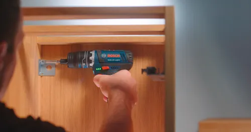 A person using a Bosch GSR12V-140FC cordless drill to install a metal bracket onto a wooden cabinet.