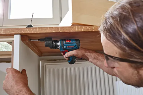 Man uses a Bosch GSR12V-140FC drill to screw a screw into a wooden surface.
