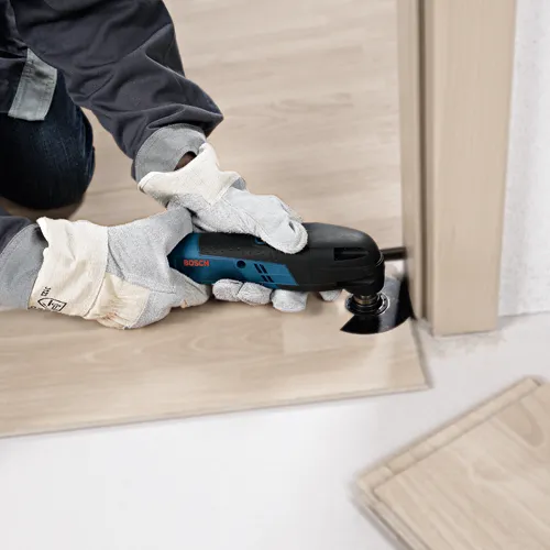 Person wearing gloves using a BOSCH multi-tool to cut flooring next to a door frame.