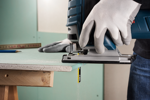 A person wearing gloves uses a Bosch jigsaw to cut a piece of drywall; a ruler and pencil are visible on the drywall surface.