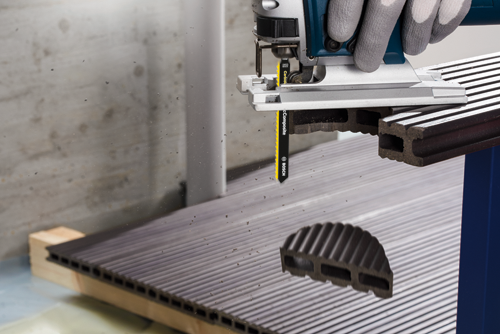 Bosch jigsaw cutting composite material with visible "Carbide" blade, "Swiss Made" printed on the blade. Hand wearing grey gloves.