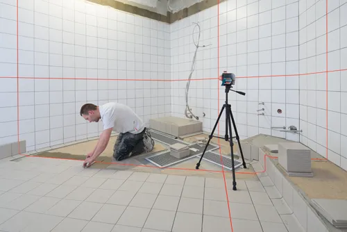 A person wearing a white t-shirt and dark pants kneels in a bathroom laying tile, a Bosch laser level on a tripod emits red lines, guiding the tile work.