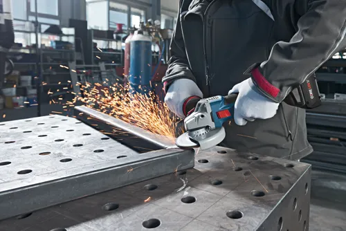 A person grinding metal with a Bosch CORE18V 6.3 Ah angle grinder, sparks flying.
