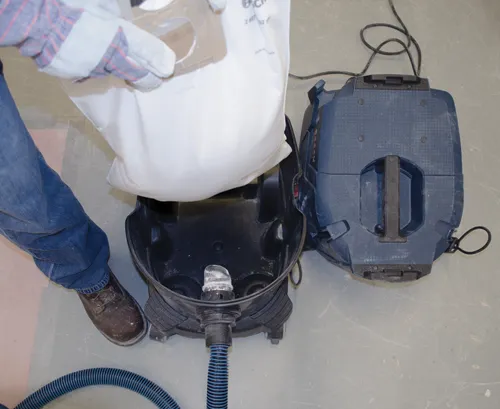 A person wearing jeans, work gloves, and brown work boots pours from a white bag into a Bosch vacuum cleaner.