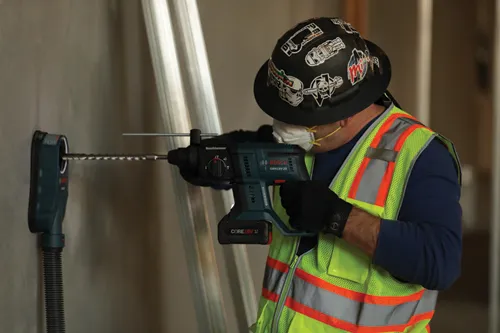 Person wearing a hard hat and safety vest using a Bosch GBH18V-20 drill to bore a hole into a wall.