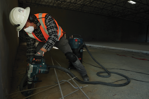 Person wearing safety gear using a Bosch 11264EVS hammer drill with a vacuum nearby.