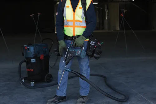 Person wearing work attire holding a drill and standing in front of a Bosch HEPA Ready vacuum.