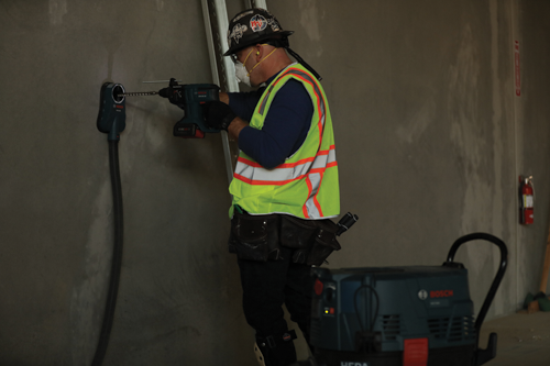 Person drilling into wall with Bosch GSB18V-20 drill while connected to Bosch dust extraction system, wearing safety vest.