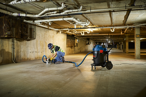 Person wearing safety gear cutting concrete floor, a Bosch HEPA Ready vacuum cleaner nearby. Building has pipes and a support column.