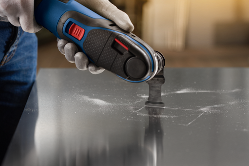 A person wearing gloves using an oscillating multi-tool to cut into a dark surface, creating a dust trail.