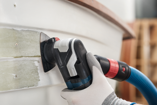 Bosch StarlockMax multi-tool being used on a white surface, with a gloved hand holding the tool, a blue hose, and the word "Bosch" visible on the tool.