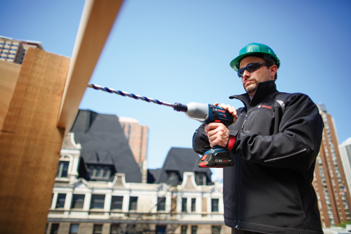 A person wearing a green helmet and sunglasses drills into wood with a Bosch drill. The drill has the Bosch logo and "CORE18V" printed on it. The person is wearing a black jacket with the Bosch logo. Buildings and a clear blue sky are in the background.