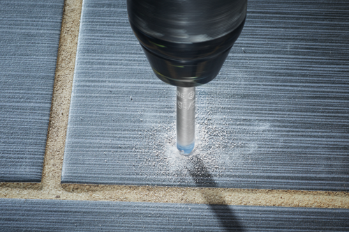 Drill bit drilling into gray tile, generating dust. Some text is visible on the drill bit.