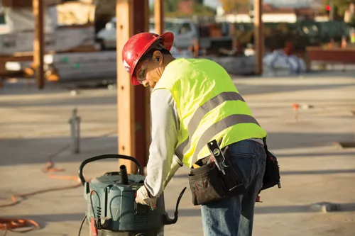 Construction worker wearing a red hard hat and yellow safety vest, inspecting a vacuum cleaner.