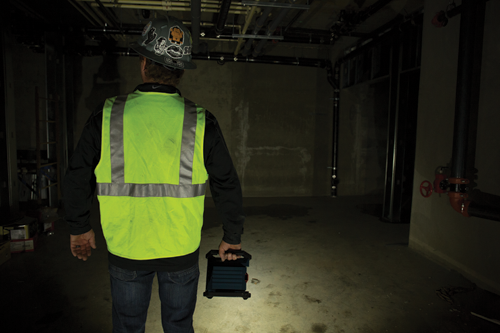 Person in fluorescent safety vest and hard hat holding a work light in a dimly lit unfinished interior.