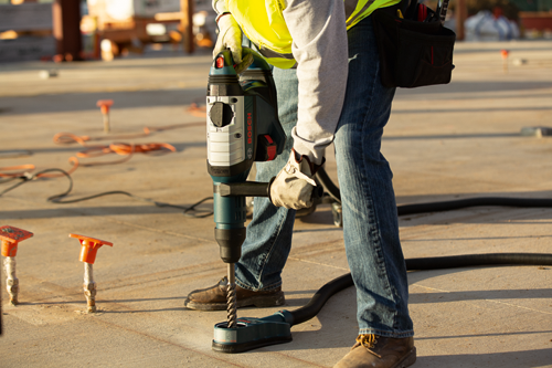 Person holding Bosch rotary hammer drill, drilling into concrete. The tool is connected to a vacuum hose. The person wears jeans, a yellow safety vest, and gloves.