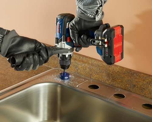 Bosch GSR18V-755C drill, drilling a hole in a stainless steel sink with a blue EDGE hole saw bit; gloved hands visible.