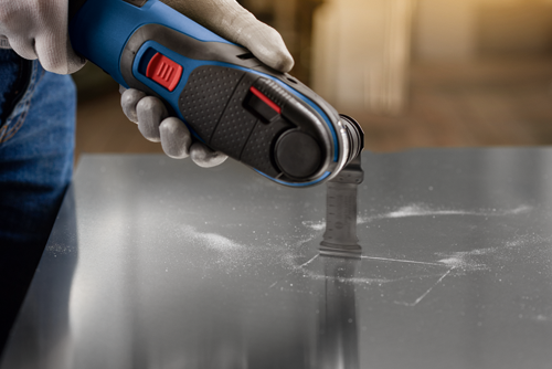 A person wearing gloves uses a blue and black power tool to cut into a gray surface, creating a line and dust.