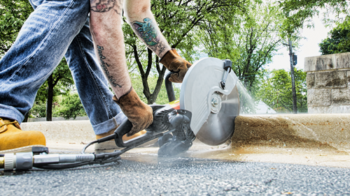 Person wearing work gloves and jeans uses a circular saw to cut a concrete edge; tattoos are visible on the person’s arm.