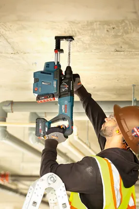 Bosch GBH18V-26D Bulldog rotary hammer being used by a person wearing safety glasses, earplugs, a hard hat, and gloves to drill into a ceiling.