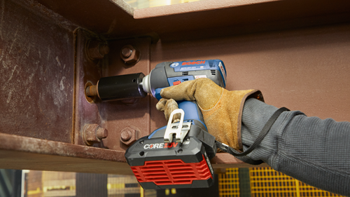 Bosch GDS18V-221 cordless impact wrench, with a black socket, is held by a person wearing a brown glove. The wrench is mounted to a rusted metal structure, with bolts.