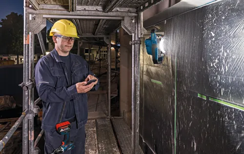 Person wearing a yellow hard hat and glasses holding a phone, on scaffolding near a Bosch LED work light and Bosch drill.