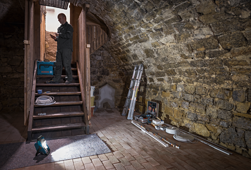 A person stands on a staircase holding a smartphone near a Bosch toolbox and a floodlight in a basement. Additional tools and a ladder are present.