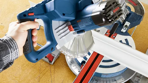 Bosch miter saw cutting white trim, held by a person with a plaid shirt sleeve visible.