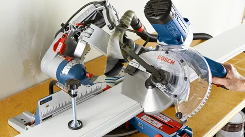 Bosch miter saw cutting white wooden piece, visible "BOSCH" on blade guard and body.