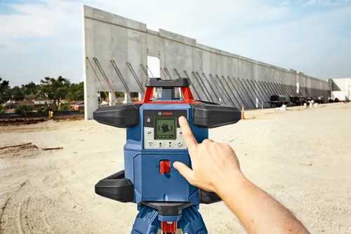 Bosch laser level, a person's hand pressing a button on the device. A construction site is in the background.
