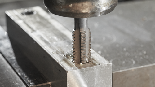 Metal tap tool cutting into a metal block, shavings visible.