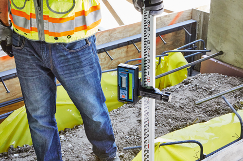 A person wearing a high-visibility vest and jeans holding a Bosch measuring tool next to a measuring rod.