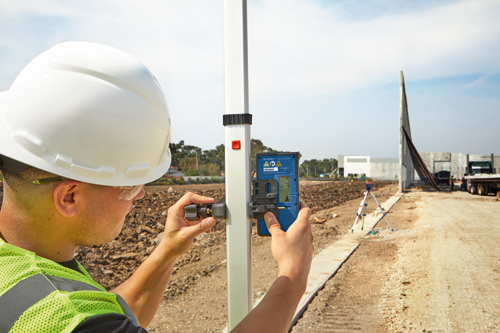 A person wearing a white hard hat and a safety vest is holding a blue measuring device, model LRD 360, on a white pole, in a construction site.